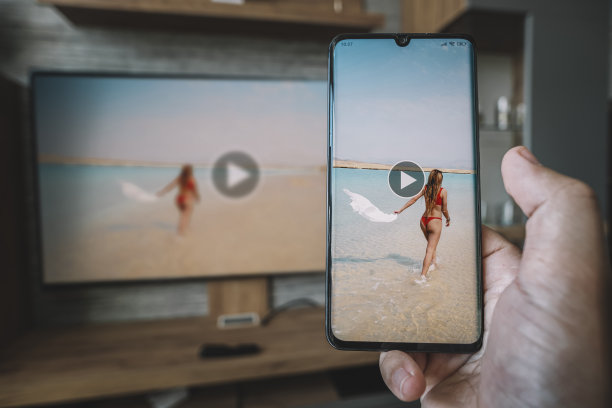 拿着智能手机的男性的手，对面是有大屏幕电视的客厅。使用wi - fi传输图像从手机上的显示器屏幕。在电视上观看智能手机上的视频。图片下载