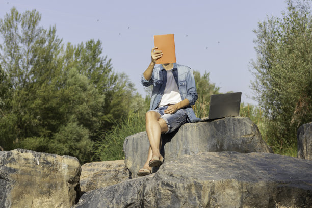 一名男子手持一本杂志，旁边是他的笔记本电脑，坐在自然界的一块岩石上图片下载