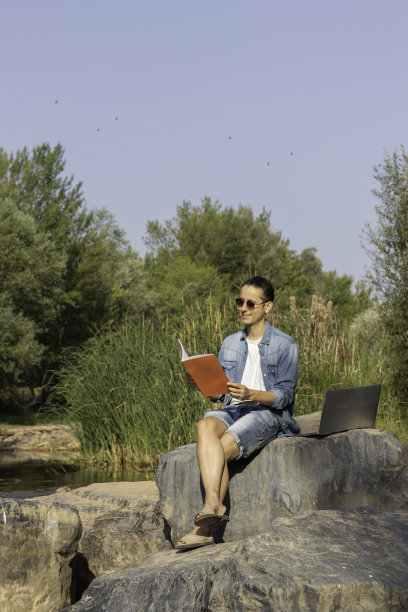 数码游牧民坐在河边的森林里阅读和工作的垂直照片图片下载