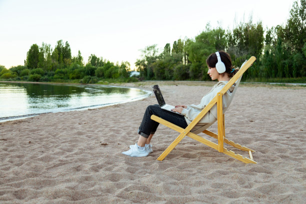 戴着耳机的自由职业女性斜坐在湖边用笔记本电脑工作图片下载