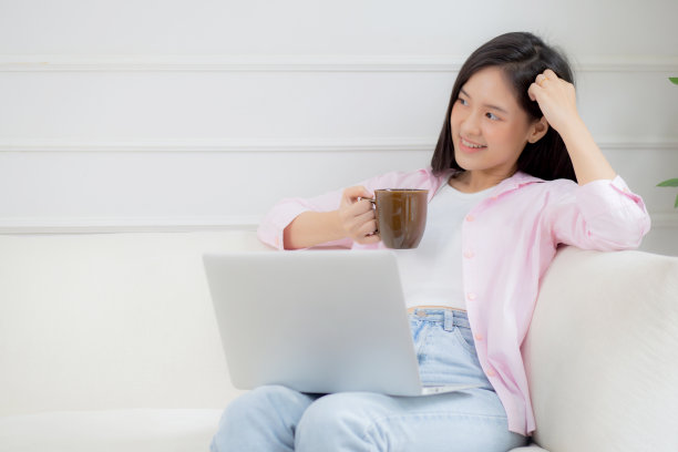 年轻的亚洲商业女性在家工作，带着笔记本电脑，在客厅的沙发上喝咖啡，自由职业的女孩用笔记本坐在沙发上舒适放松，生活方式的概念。图片下载