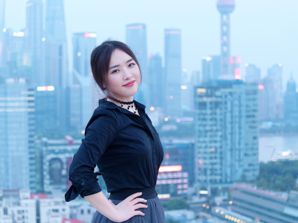 美丽的年轻中国女人在大厦屋顶上的黑色摆拍与模糊的上海外滩地标建筑背景与黄昏的光。情感、人、美和生活方式理念。图片下载