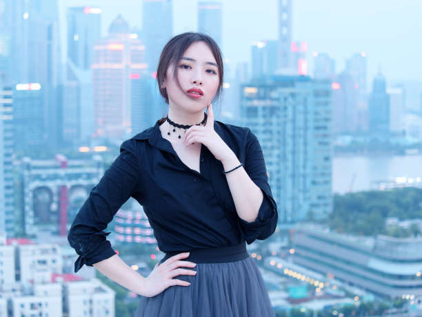 美丽的年轻中国女人在大厦屋顶上的黑色摆拍与模糊的上海外滩地标建筑背景与黄昏的光。情感、人、美和生活方式理念。图片下载