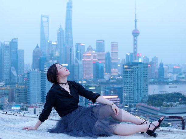 美丽的年轻中国女人坐在大厦的屋顶上，模糊的上海外滩地标建筑背景与黄昏的光。情感、人、美和生活方式理念。图片下载