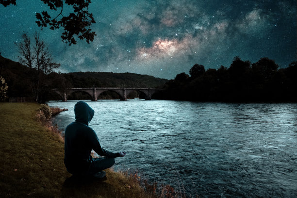 夜晚，一个人坐在河岸边冥想的剪影，背景是银河系图片下载