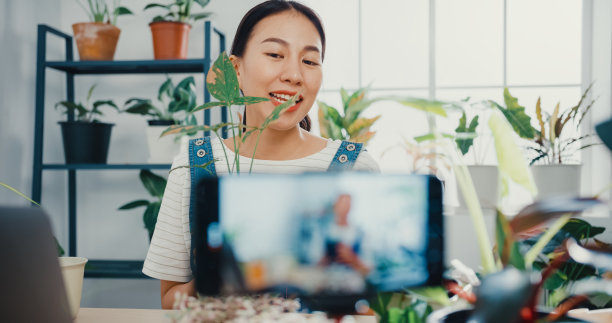 一名年轻的亚洲女性在树木商店用手提电脑接收订单，并在早上通过电话直播植物录制视频。图片下载