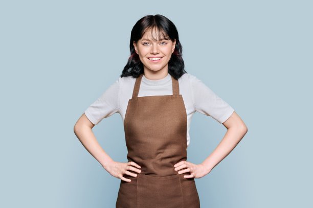 微笑的年轻女性员工在围裙看着相机在灰色孤立的背景图片下载