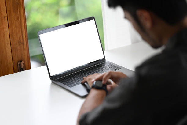 年轻男员工坐在办公桌前的笔记本电脑前，用智能手表查看时间。在肩膀的观点。图片下载