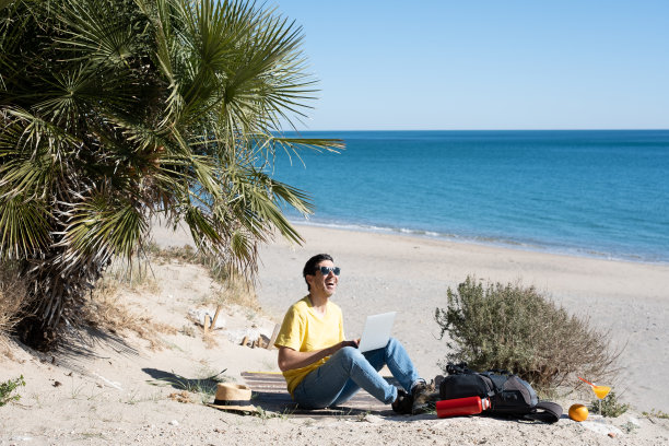 一名年轻人在暑假期间的旅行中，在海滨海滩上打理他的数字业务。在旅行中工作的概念。图片下载
