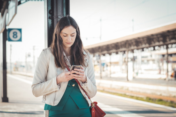 微笑的商业女性在火车站等待时使用智能手机-迷人的年轻学生女孩站在火车站站台上使用手机图片下载