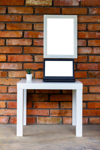 一台黑色笔记本电脑和一个白色的模型放在一张白色的桌子上，靠着一堵旧砖墙，白色的模型放在一个白色的相框里图片下载