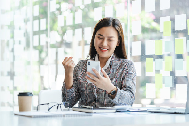 兴奋的亚洲年轻女子坐在桌子上使用电话感到高兴。图片下载