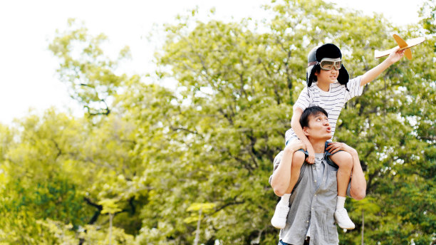 亚洲家庭生活方式父子俩坐在他的肩膀上在公园里跑步。纸飞机作为孩子手中的玩具和有许多大树在花园里是自然的夏天的早晨图片下载