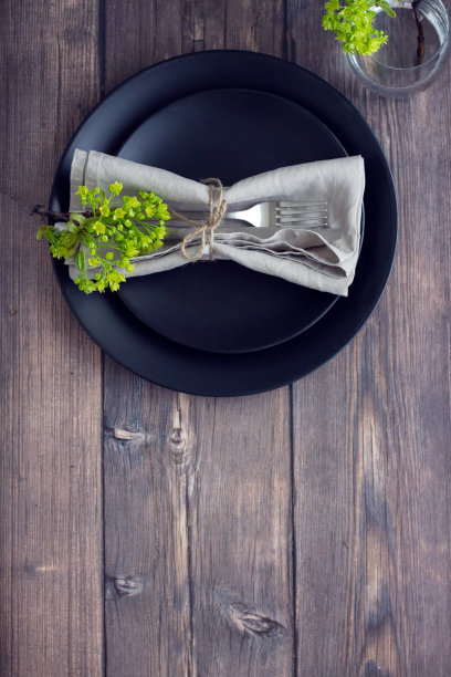 木制的桌子上有黑色的盘子，银色的餐具，一杯水和亚麻餐巾。餐厅设置。图片下载