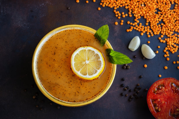 俯视图的传统土耳其扁豆汤与柠檬和薄荷在陶瓷碗，堆红扁豆，黑胡椒子，蒜瓣和减半的西红柿在黑暗的背景。低调照片。有选择性的重点。模糊的背景。图片下载