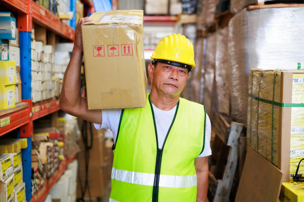 在工业仓库工厂的货架上搬运产品纸板箱的亚洲老年男性仓库工人。黄色安全安全帽头盔。图片下载