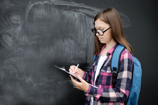 十几岁的学生女孩，衬衫，眼镜，蓝色背包和日记或笔记本在黑色的黑板背景。教育、学校和人的观念。十几岁的女孩在黑板旁边看书。副本的空间。图片下载