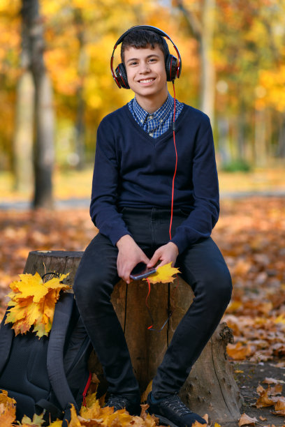 一个十几岁的男孩坐在秋天的公园里，戴着耳机听音乐，在一个明亮的阳光灿烂的日子里，树上有黄色的叶子图片下载