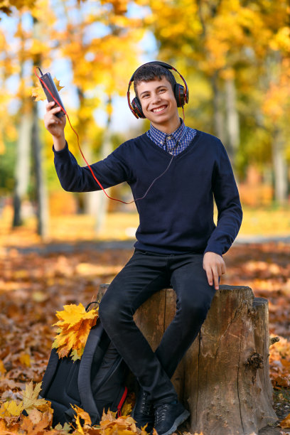 一个十几岁的男孩坐在秋天的公园里，戴着耳机听音乐，在一个明亮的阳光灿烂的日子里，树上有黄色的叶子图片下载