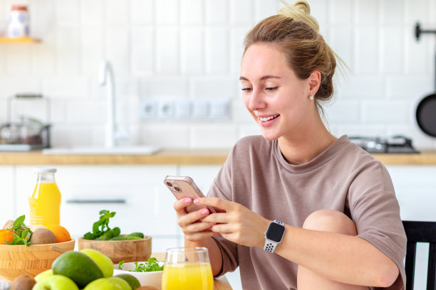 健康的生活方式。美丽快乐的女人早餐有健康的食物，沙拉和新鲜的果汁。放松，坐在厨房用手机聊天，友好的微笑图片下载