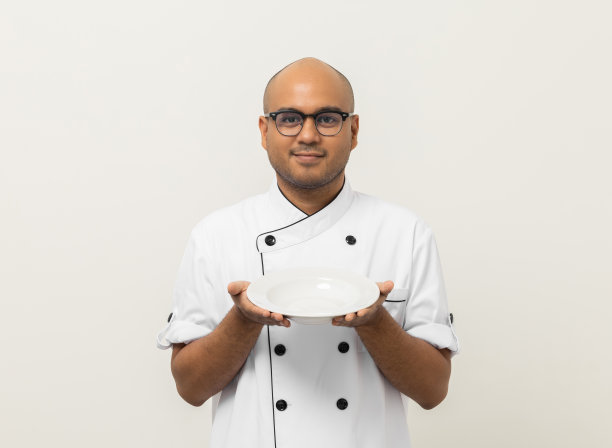微笑年轻英俊的亚洲男子厨师在制服拿着空盘子美味的菜菜单好味道上孤立。烹饪印度人职业厨师或面包师，厨房、餐厅和酒店的人。图片下载
