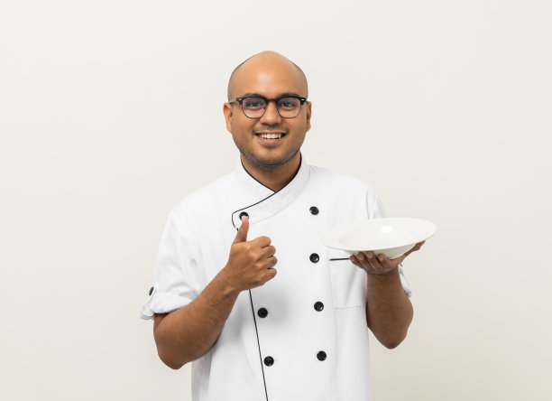 微笑年轻英俊的亚洲男子厨师在制服拿着空盘子美味的菜菜单好味道上孤立。烹饪印度人职业厨师或面包师，厨房、餐厅和酒店的人。图片下载