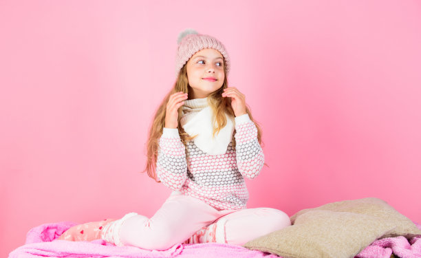 冬季配件的概念。女孩长发梦幻的心情粉红的背景。孩子微笑着穿针织配饰。小女孩戴着可爱的针织时尚帽子和围巾配饰。冬天的时尚配件图片下载