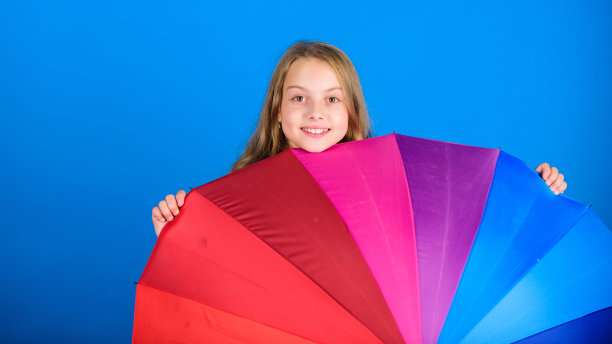 点亮生活。小孩从五颜六色的彩虹伞里探出头来。颜色你的生活。女孩兴高采烈地躲在伞后。五彩缤纷的雨伞配件。天气预报的概念。雨天也要保持积极图片下载