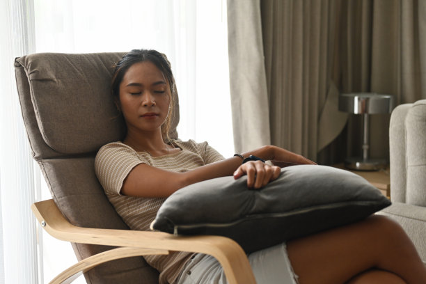 亚洲古铜色皮肤女性的肖像在扶手椅上打盹，拿着一个枕头，在现代的房子内部装饰。家和生活的概念。图片下载