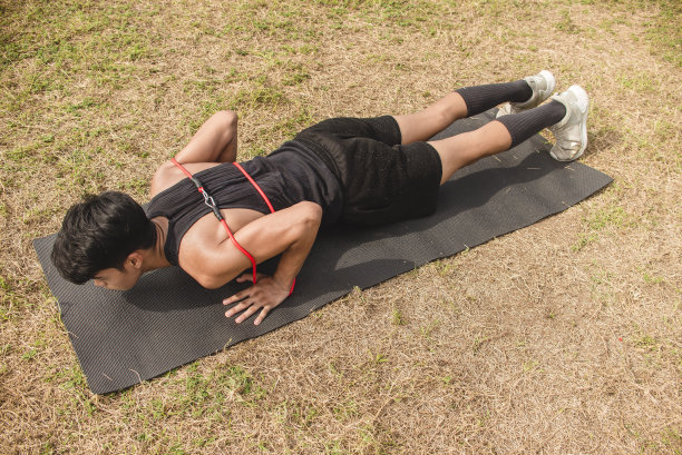 一名年轻的亚洲男子在户外的一块黑色垫子上做一组带着阻力带的俯卧撑。胸部或全身锻炼。图片下载