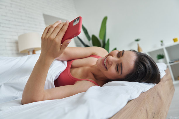 迷人的年轻女子使用智能手机和微笑，而躺在家里的床上图片下载