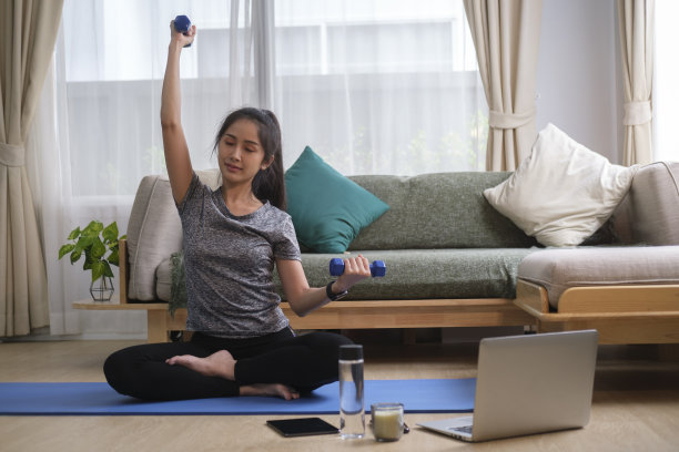 运动的年轻女性在笔记本电脑上看辅导课，在家用哑铃锻炼。图片下载