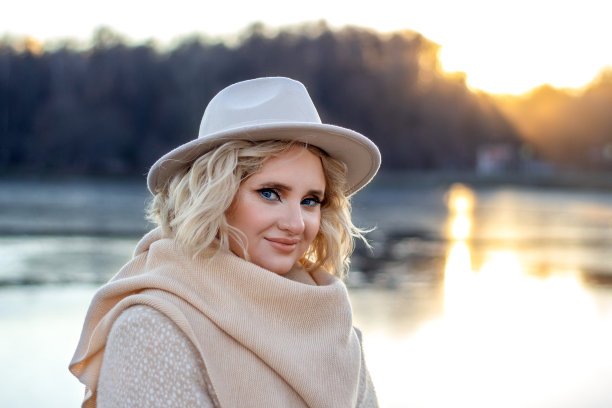 一个戴着帽子的金发女人的肖像，在秋天的夕阳下，在自然的背景下——水和森林中，画着明亮的眼妆。图片下载