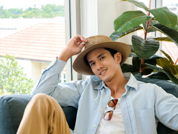 帅气的亚洲男人穿着浅蓝色牛仔衬衫和白色t恤，戴着墨镜和棕色沙滩帽，坐在舒适的沙发座位上，靠近窗户和绿色的叶子在客厅里，微笑着看着镜头。图片下载