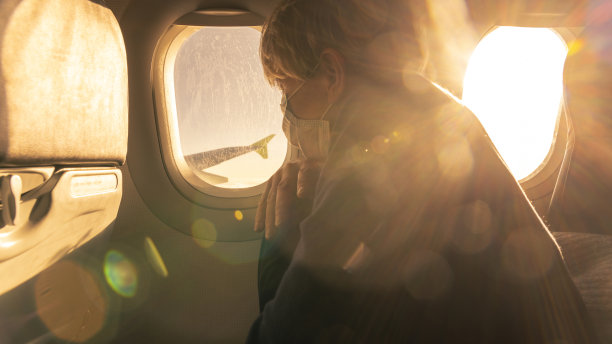 旅行期间大流行。成熟的女人戴着口罩，坐在飞机上，望着窗外。阳光会产生强烈的透镜耀斑。图片下载