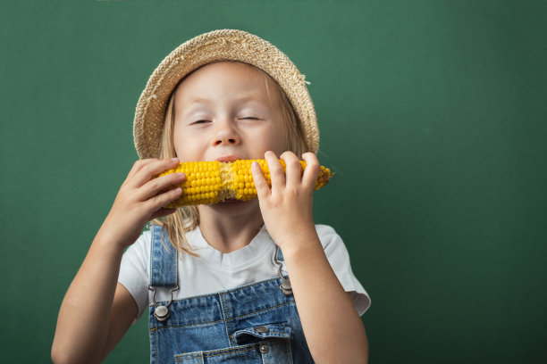 可爱的白人小女孩微笑着享受新鲜的烤玉米在深绿色的背景。快乐蹒跚学步的女孩戴着草帽和牛仔工装裤。全国玉米棒日快乐图片下载