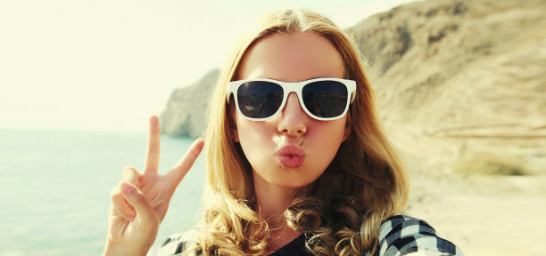 肖像特写的快乐的年轻女子吹她的嘴唇发送甜蜜的空气吻伸展手，以海洋为背景自拍图片下载