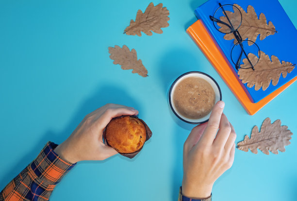 女人的手与一杯咖啡，书和蛋糕在秋天的蓝色背景图片下载