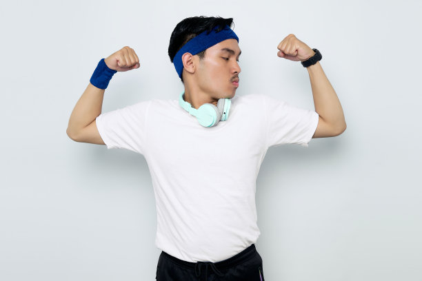 这位骄傲的年轻亚洲运动员戴着耳机，戴着蓝色头带和白色t恤，在白色背景的健身房训练后举起手臂展示肱二头肌，展示她的成就图片下载