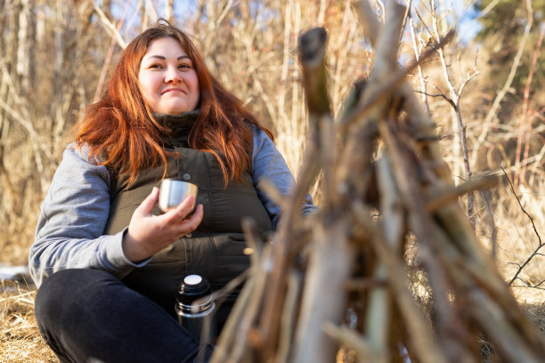 一个快乐快乐的女人在大自然休息的特写照片，盘腿坐在篝火附近拿着一杯热茶和保温瓶下来。图片下载