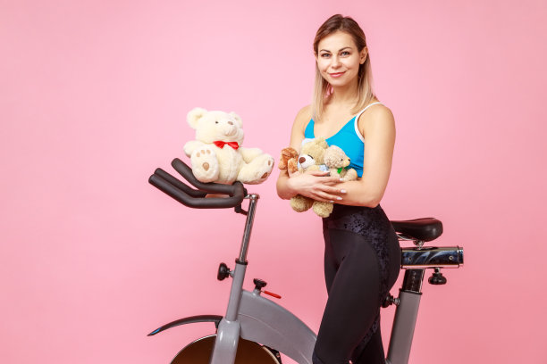 迷人的美丽运动女人抱着泰迪熊，摆姿势附近的运动设备，锻炼自行车。图片下载