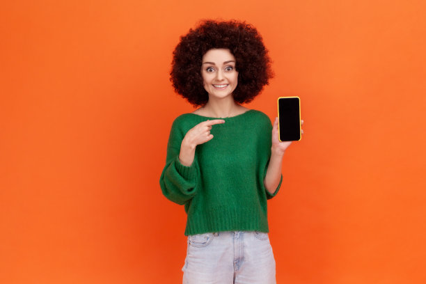笑容积极的女性，留着非洲式发型，穿着绿色休闲风格的毛衣，展示黑色黑屏的智能手机，手指指向显示屏。图片下载