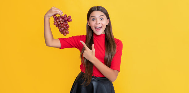 惊讶的十几岁的孩子拿着一串葡萄在黄色的背景图片下载