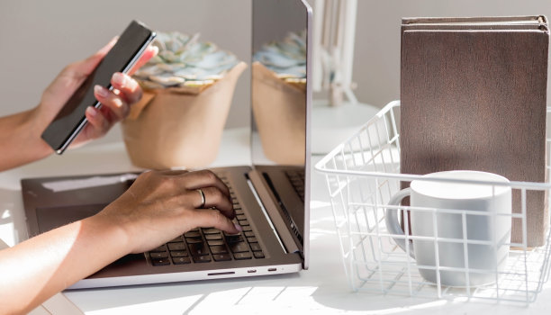 一个自由职业女性的手坐在家里的办公室里，拿着笔记本电脑和智能手机。女人在家工作。工作日的开始。人力资源管理与招聘的概念图片下载