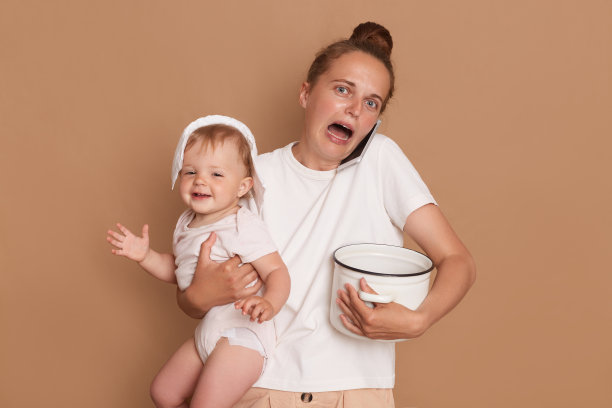 室内拍摄了一个震惊绝望的女人，她梳着发髻，穿着白色t恤，和女儿站在一起，拿着装满食物的锅，说着智能手机，尖叫，被隔离在棕色背景下。图片下载