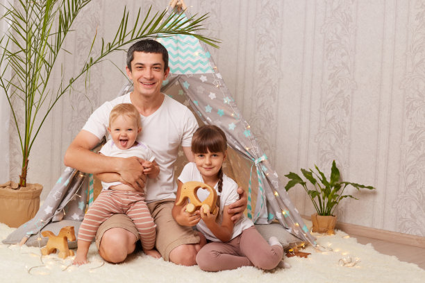 一名身穿白色t恤的男子带着孩子和大女儿在家里的儿童房棚屋附近，微笑着看着镜头，表达积极的情绪。图片下载