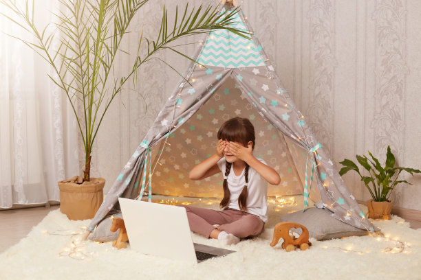 室内拍摄的小学龄前女孩坐在棚屋前的笔记本，有视频通话，用手掌遮住眼睛，隐藏自己，在新的小帐篷玩。图片下载