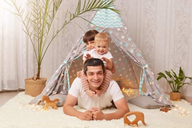 有吸引力的乐观的男人穿着白色t恤躺在小帐篷的地板上，孩子们坐在他的背上，家人看着镜头，一起度过周末。图片下载