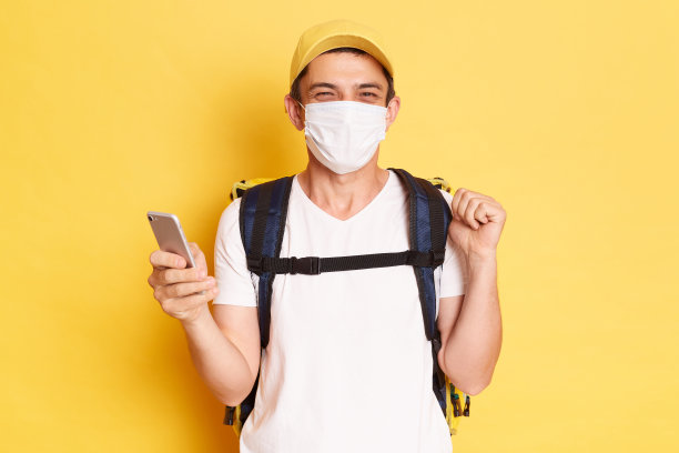 照片中，快递员背着热背包，穿着白色t恤，头戴帽子，站在黄色的墙上，拿着智能手机，看着相机，正在送餐。图片下载