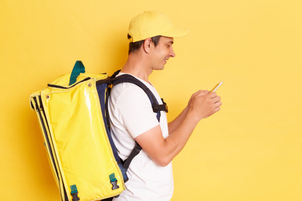 侧面肖像积极英俊的送货员与热双肩包，白色t恤和帽子站在黄色的墙，站在使用手机，与客户聊天。图片下载